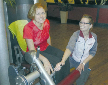 Elke Arzner sitzt auf einem Trainingsgerät für die Beinmuskulatur und erhält Erklärungen von ihrer Trainierin